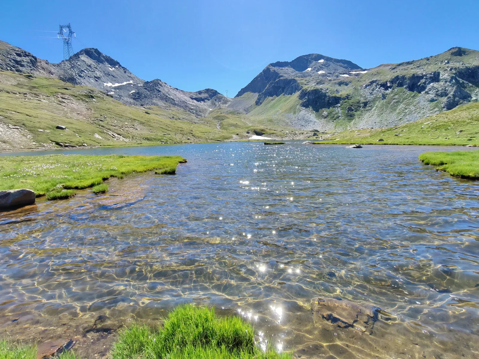 Il Lago Nero: acqua bassissima e trasparente sul fondo sassoso, con le lingue erbose che entrano nell'acqua e i tralicci sulla cresta