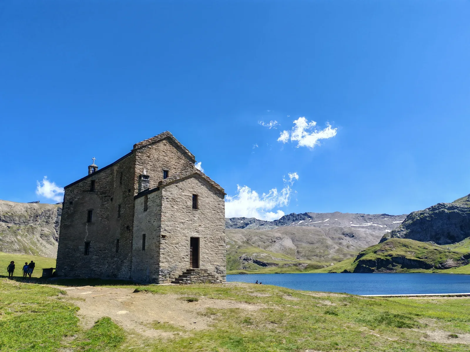 Il santuario della Madonna delle Nevi visto di fronte: l'alto edificio in pietra con il campanile a vela e la croce, il lago Miserin sulla destra e le creste della conca dietro