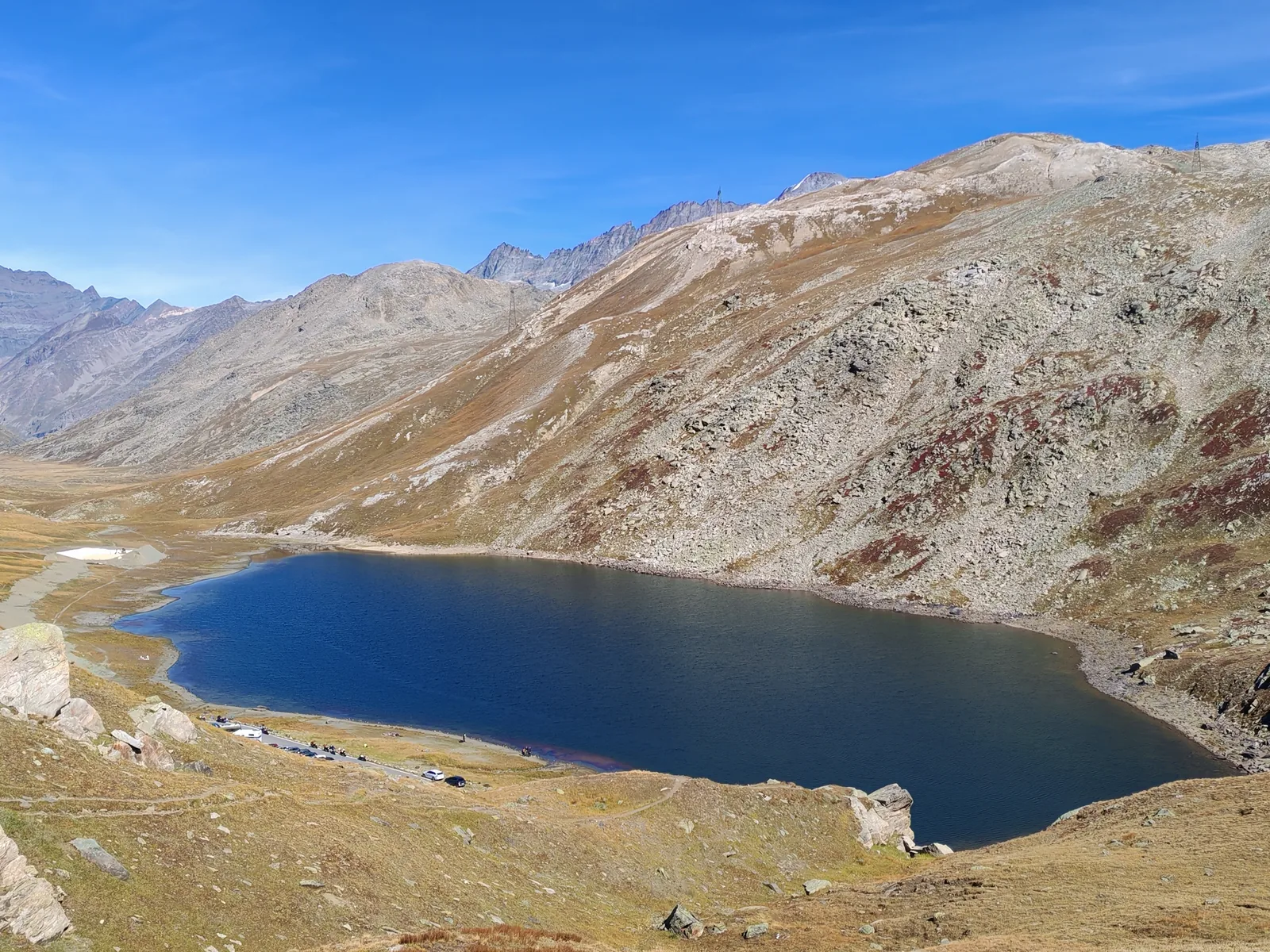 Il Lago del Nivolet visto dall'alto con la strada e le auto parcheggiate sulla riva, e la valle che si apre verso ovest