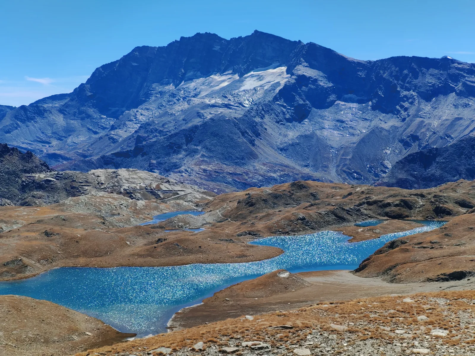 I laghi Rosset e Leità visti dal dosso a 2.828 m, due specchi d'acqua azzurri incassati fra dossi ocra, con un massiccio scuro e innevato sullo sfondo