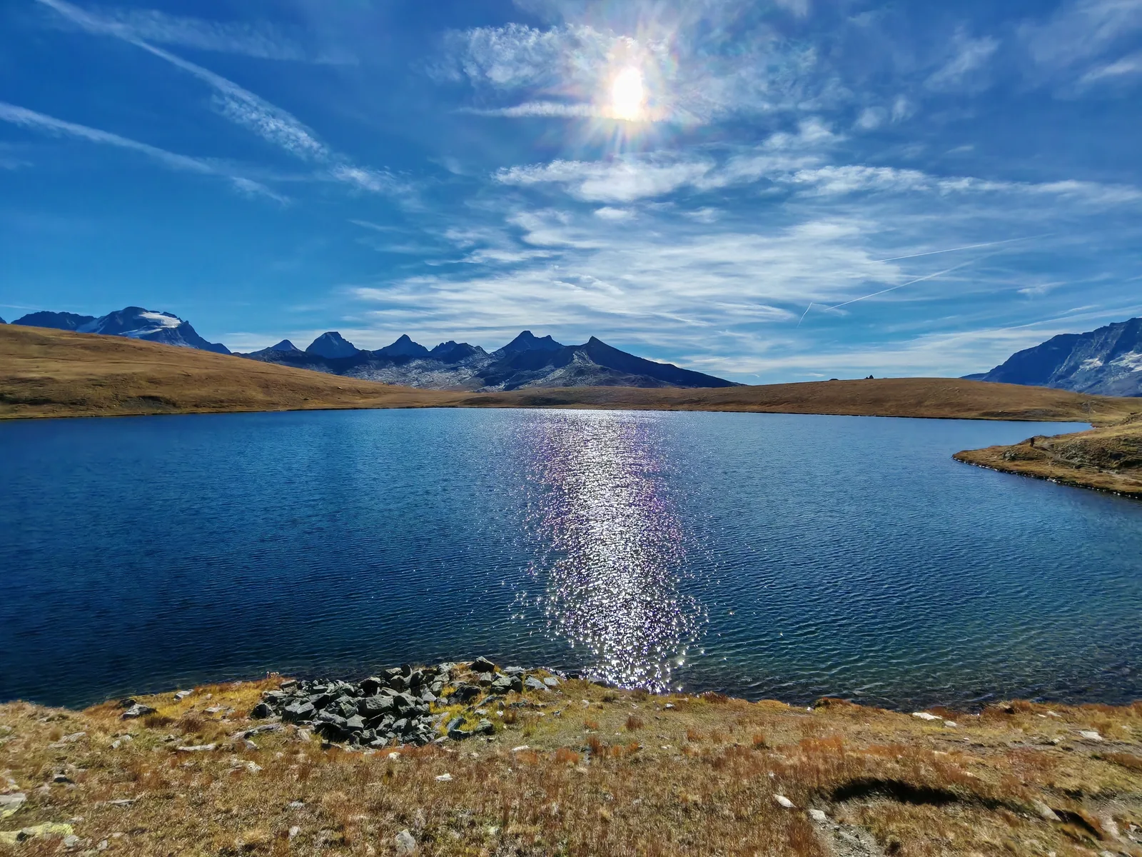 Il Lago Rosset con il riflesso del sole sull'acqua, le praterie ocra attorno e la corona di cime rocciose sullo sfondo