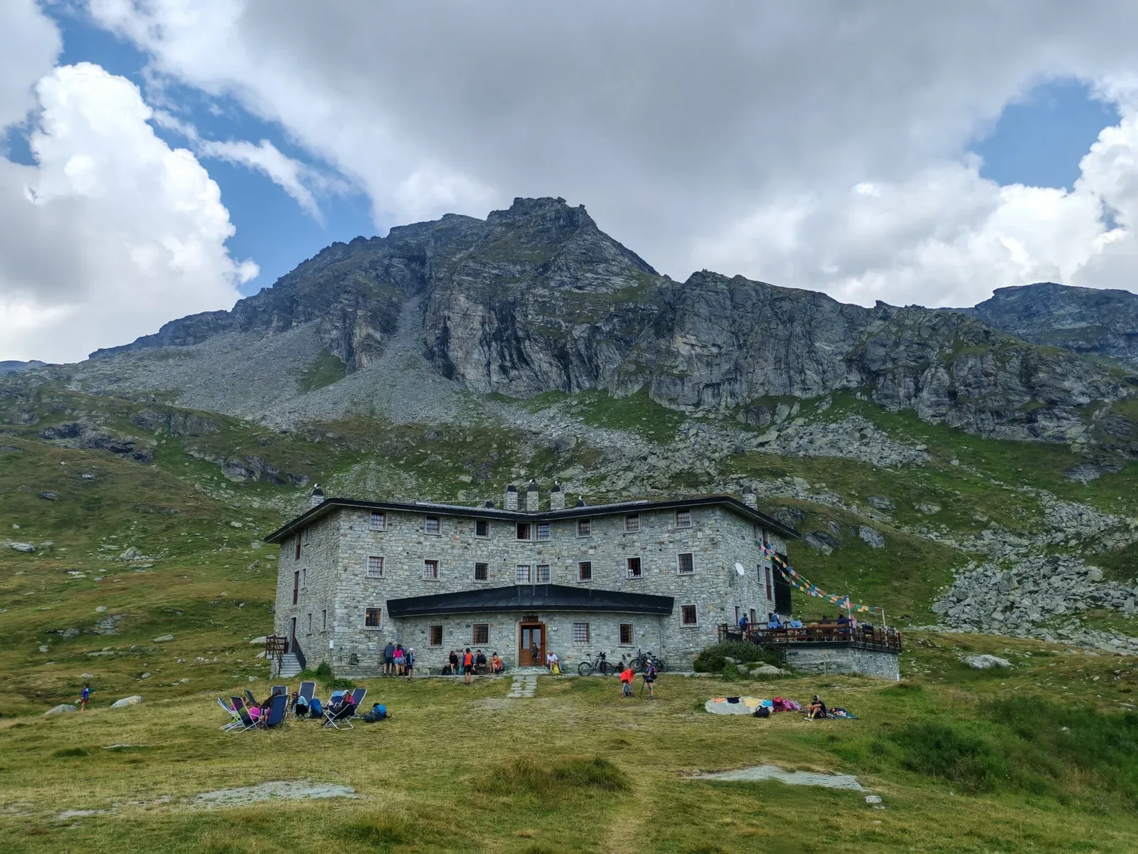 Il Rifugio Arp, grande edificio in pietra a tre piani, sotto la bastionata rocciosa, con escursionisti sul prato