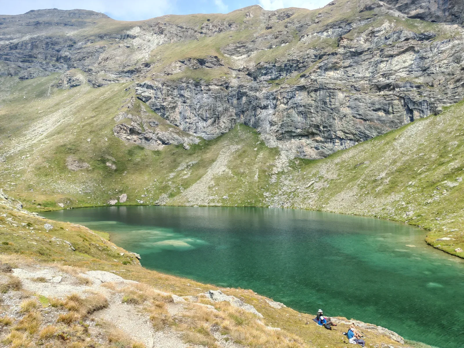 Il Lac Long dalle acque verde smeraldo sotto un'alta bastionata rocciosa, con escursionisti seduti sulla riva
