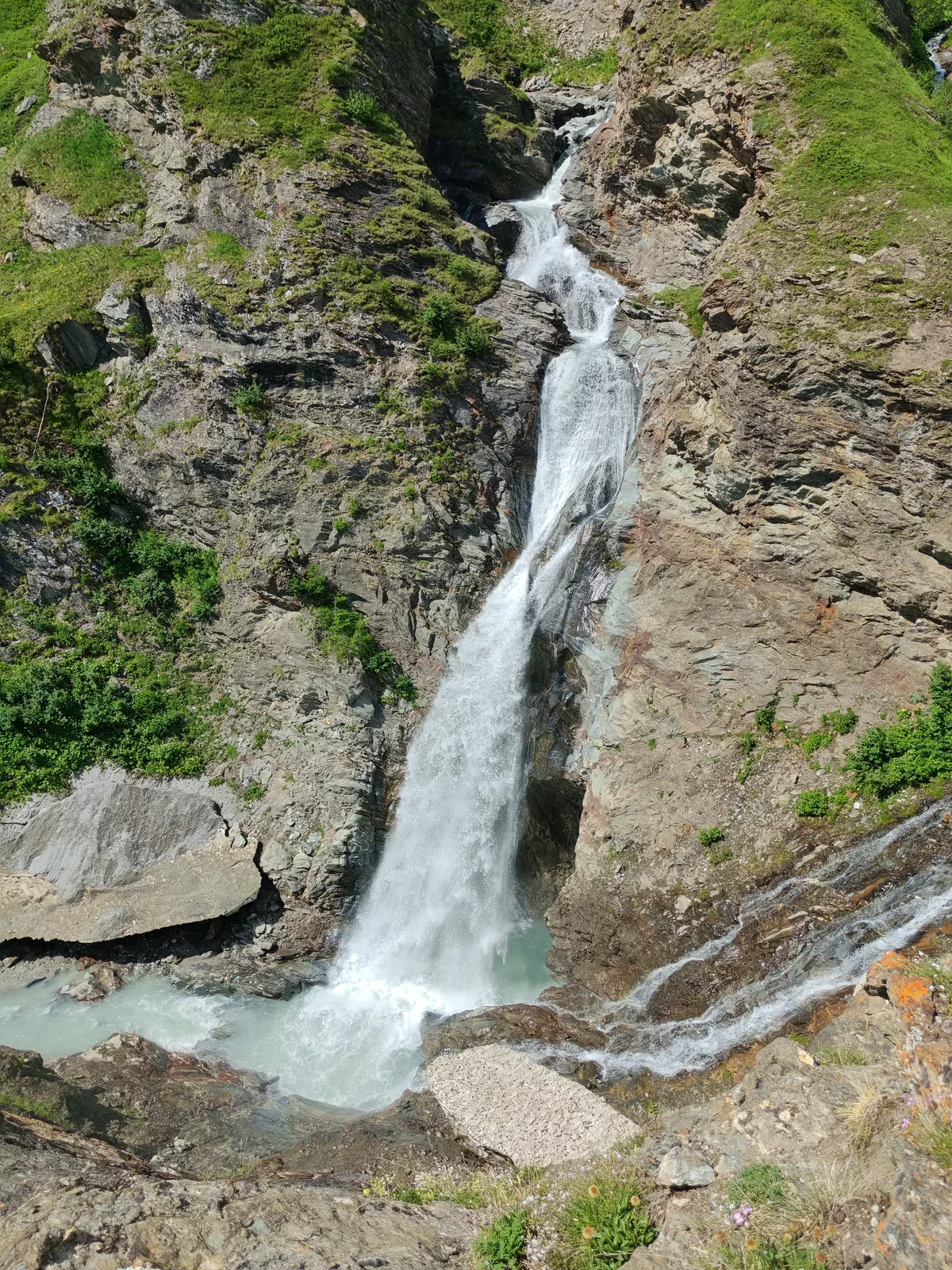 Una cascata vista dall'alto: l'acqua scende su una placca rocciosa inclinata e finisce in una pozza glaciale color verde ghiaccio