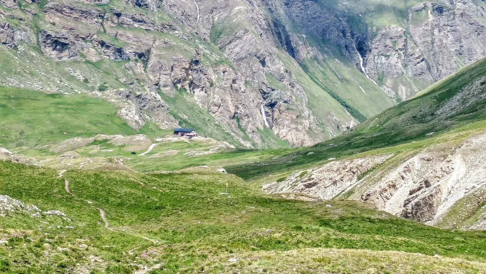 Il Rifugio Benevolo visto da lontano dal belvedere: l'edificio isolato sul ripiano erboso, con le pareti rocciose e le cascate della testata dietro