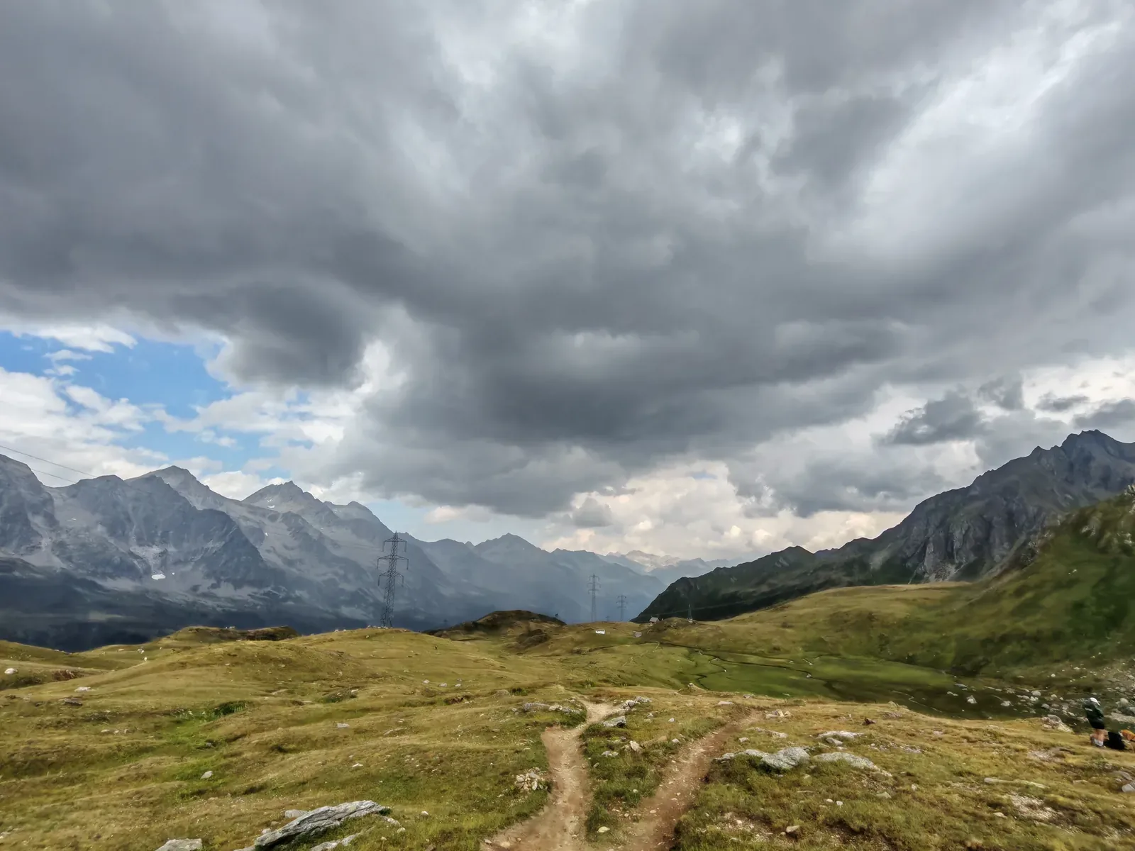 Il valico del Passo San Giacomo con il sentiero, i tralicci dell'alta tensione e le cime svizzere sotto un cielo di tempesta
