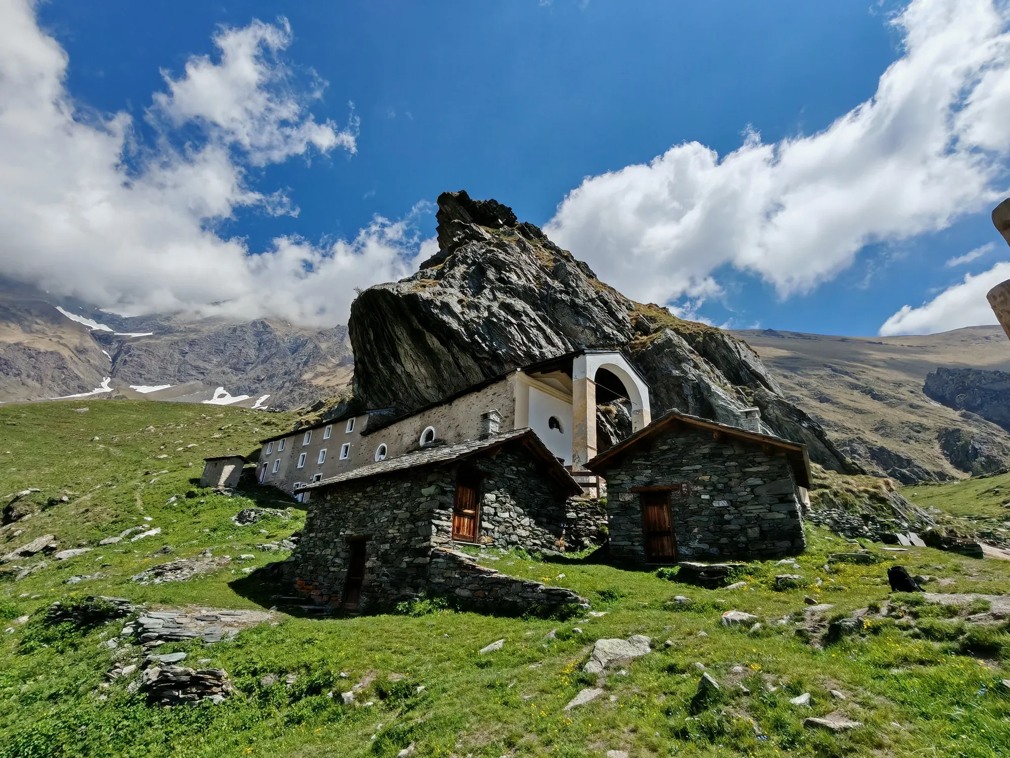 Il santuario di San Besso addossato al monolito del Monte Fautenio, con le costruzioni in pietra e lastre d'ardesia sul prato e i nevai residui sulle creste dietro
