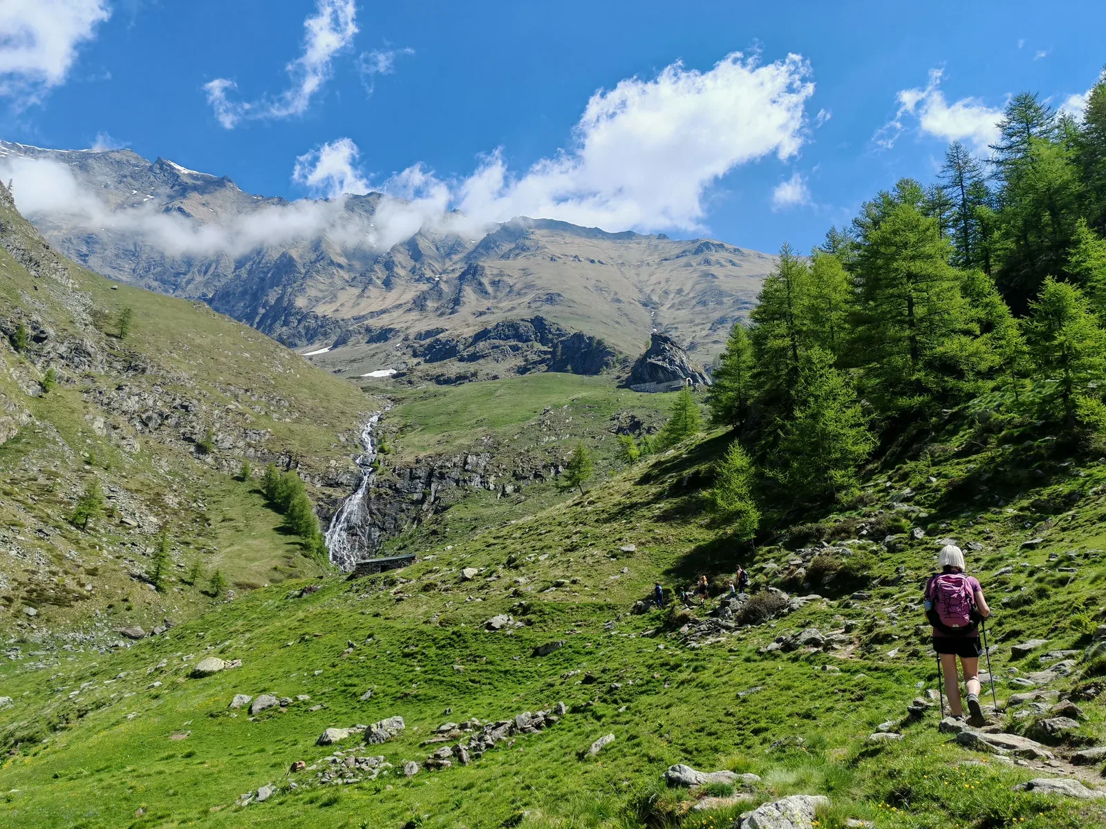 La salita nell'alta valle: escursionisti sul sentiero fra i prati, la cascata che scende dalle balze rocciose e, sul ripiano in alto, il santuario già visibile